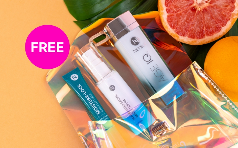 Neora’s Summer Skin Essentials Set, which includes two Age IQ Day Cream, Moisture-Lock Lip Mask and Hydrating Mist with a FREE Holographic Travel Bag lying on a lead and some grapefruit